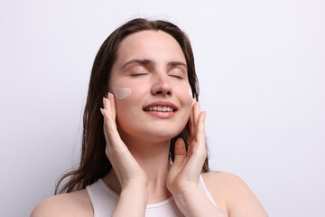 Smiling woman applying skincare product onto her face on light grey background, closeup