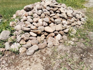 A pile of round or oval river rocks that looks quite high is placed on the surface of open ground that is overgrown with green weeds, and there are small shadows between the pile of rocks.