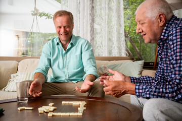 A grown son and his elderly father enjoy a quiet game of dominoes in the living room, sharing connection, laughter, and meaningful time together.