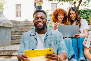 Diverse university students sitting on campus stairs, working on digital tablets and laptops, engaging in collaborative learning through technology