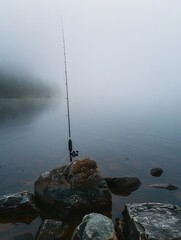 Serene fishing scene at dawn with a lone fishing rod on a rocky shore amidst a foggy lake