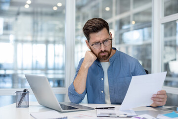 Naklejka premium A businessman in glasses reviews paperwork at his desk in a modern office setting, with a laptop and thoughtful expression.