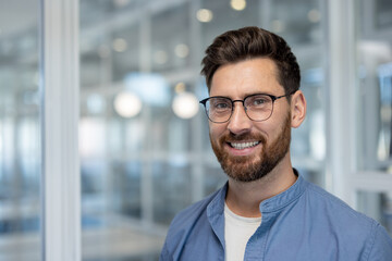 Obraz premium A smiling man with glasses and a beard poses for a portrait in a modern office environment.