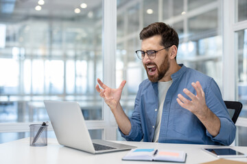 An exasperated businessman, wearing glasses, shouts in frustration while working on his laptop at...