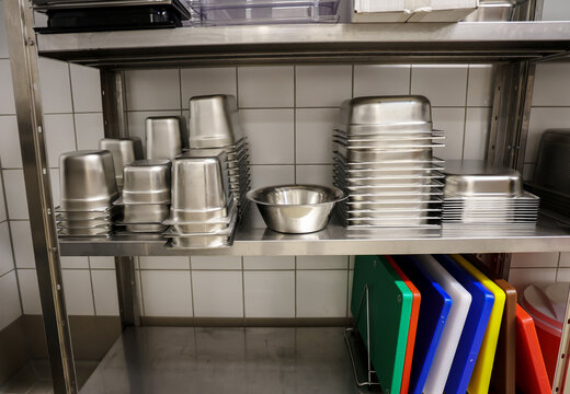 Stacked stainless steel containers and mixing bowls on a clean metal shelf in a commercial kitchen. Below, organized color-coded cutting boards for safe food preparation.