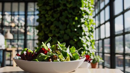 Mixed green salad in white bowl sits on table in contemporary restaurant with large windows and vertical garden wall, creating a fresh and inviting atmosphere - Powered by Adobe