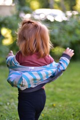 Obraz premium Happy red hair toddler girl running outdoors on a sunny day with arms outstretched, holding a striped jacket. Carefree childhood moment captured in a vibrant green garden with warm natural light.