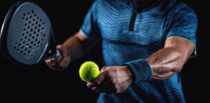 Padel player in action preparing to serve, holding paddle and bright tennis ball, focus on hands and equipment, intense sports moment, dark background.