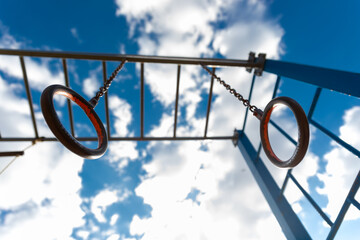Upward view of metal gymnastic rings hanging from chains, with a blue sky and white clouds in the background.
