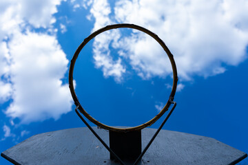 Upward view of a worn basketball hoop without a net against a vibrant blue sky with scattered clouds.