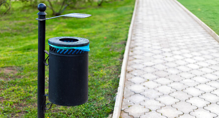  Black metal trash bin with blue liner mounted beside a patterned paved walkway in a green public...