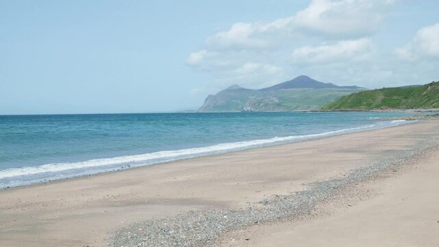 Deserted sandy beach with waves at Nefyn, Wales, UK, with a view across a bay The Rivals, Yr Eifl, mountain peaks in the distance under a blue sky with cloud.