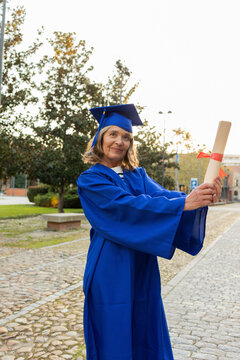 Mature woman in blue graduation gown holding diploma on campus