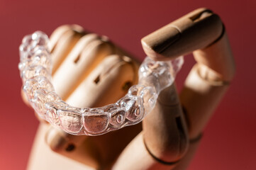Close-up of wooden elegant hand holding invisible clear aligners, on pastel pink background. Healthy lifestyle and teeth concept