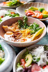A bowl of ramen with chicken, corn, mushrooms, cilantro, and a soft-boiled egg, surrounded by fresh salads and vegetables. A colorful and appetizing meal presented in a gourmet setting