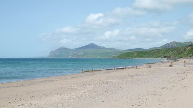 Sandy beach with groynes at Morfa Nefyn, Wales, UK, with waves lapping the shore of a bay and views across the sea to a mountain headland under a blue sky with clouds. 
