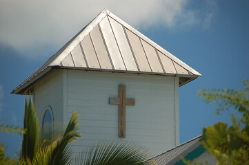 Close-up of a small white wooden church tower with a metal roof and a wooden cross, surrounded by tropical plants under a blue sky