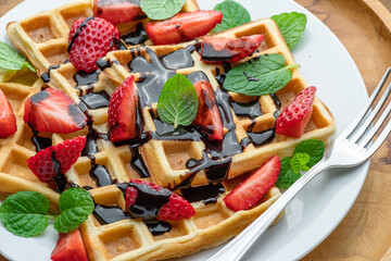 Belgian waffles with chocolate topping, strawberry slices and mint leaves on plate closeup.