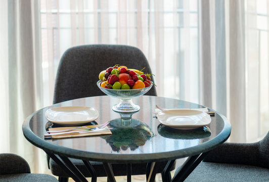 Softly lit secene of colorful fresh fruit bowl on marble tabletop with plates and chairs in the background in front of a window