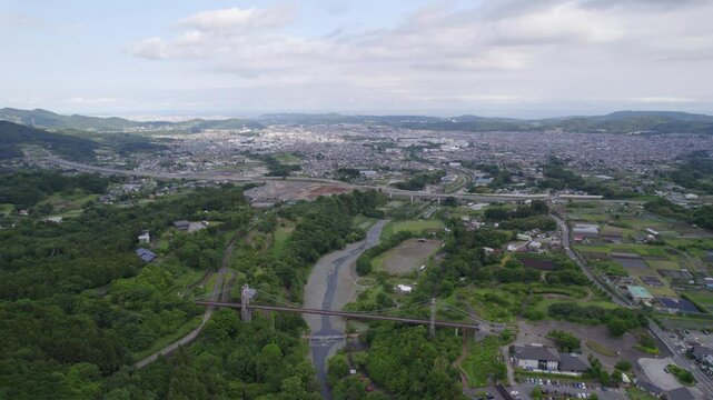 Drone view of a suspension bridge over a river in Hadano, Japan. The shot features a lush park, surrounding forest, distant mountains, and the city spreading out into the background.
