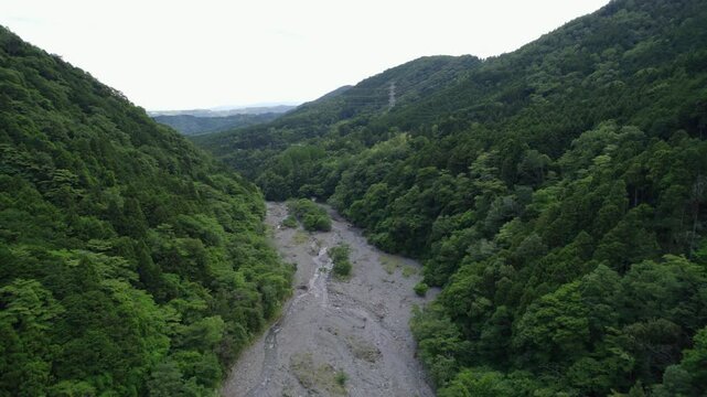 Drone view of a forested valley in Hadano, Japan, with a rocky riverbed cutting through dense green mountains. A tranquil and wild natural landscape seen from above.