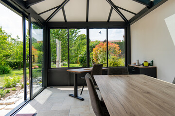 Véranda moderne lumineuse avec baies vitrées et mobilier contemporain, ouverte sur un jardin verdoyant. © Thomas