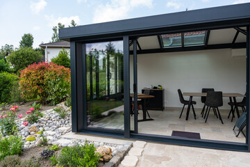 Véranda moderne lumineuse avec baies vitrées et mobilier contemporain, ouverte sur un jardin verdoyant. © Thomas