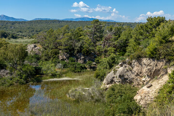 View from a hill over the natural reserve of Patara: a small lake or wetland surrounded by rocky cliffs and pine trees, with distant mountains under a blue sky.