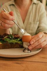 Close-up of female hands with cutlery having breakfast with egg and bacon with freshly greens served on wooden table in sunny outdoor cafe. Part of a series