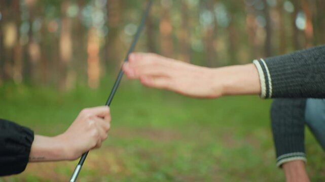 Close up of playful moment between two campers squatting in forest, one reaching to collect tent pole while other holds it back teasingly before eventually handing it over during setup process