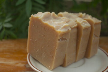 Handmade Tamarind Oatmeal Soap Bars. A stack of handmade tamarind and oatmeal soap bars placed on a white dish, photographed outdoors in natural lighting with a green garden background. 
