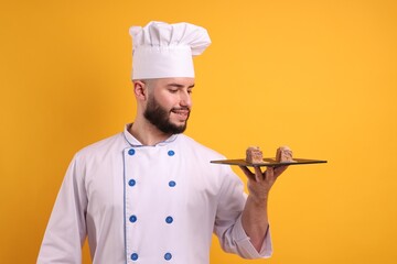 Happy confectioner in uniform holding chocolate desserts on orange background