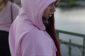 A woman in a cozy pink hoodie stands by calm water, enjoying the view. Nearby, another person...