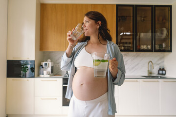 In a contemporary kitchen, a pregnant woman with a joyful expression holds a pitcher filled with...