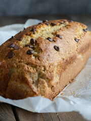 Close-up of a banana bread with chocolate chips on baking paper on a rustic wooden table. Natural light. Breakfast concept, artisanal pastries, home cooking.
