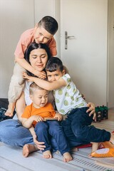 A loving mother sits on the floor, gently hugging her three young children in a bright, cozy room. The kids laugh and cuddle close, capturing warmth, care, and joyful motherhood