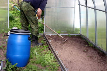 An elderly farmer rakes the soil after weeding a bed in a greenhouse. Agriculture, preparing the soil for planting plants - horizontal photo