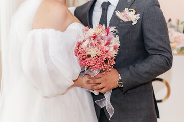 Bride and groom are holding a bouquet of flowers. The bride is wearing a white dress and the groom...