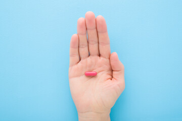 Young adult woman opened palm holding and showing red pill for improve of iron level in blood on light blue table background. Pastel color. Closeup. Top down view.