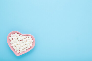 White pills in pink heart shape plate on light blue table background. Pastel color. Closeup. Healthcare concept. Empty place for text. Top down view.