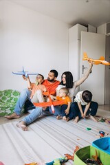 A happy family of five sits barefoot on a rug in a bright room, playing with toy airplanes and wooden trains, enjoying joyful, relaxed moments of togetherness and bonding at home.