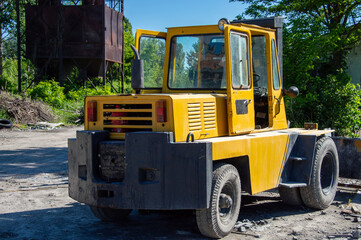 An industrial vehicle in bright yellow is parked in an outdoor location, surrounded by trees and remnants of industrial equipment