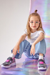 A charming girl with glasses sits on the floor in a relaxed pose. The child is dressed in a white T-shirt and blue jeans. On her feet are gray and pink sneakers with star patterns