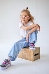 A young girl with long hair fluttering in the wind sits on a wooden cube, resting her hands on her knee and dreamily looking into the distance. The child looks relaxed and confident.