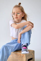 A young girl with glasses and long hair blowing in the wind, tied into a small bun on top, sits on a wooden box with a thoughtful expression on her face. The child sits with his hands folded on knees.