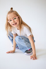 Mischievous young girl with long hair tied in a bun on top of her head sits on her haunches. Cheerful child in glasses laughs and touching the floor with her hands on a minimalist white background