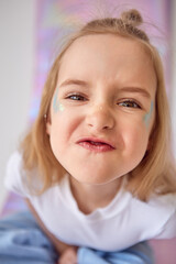 A little cute girl is making a funny face. The child has a mischievous expression, her eyes are squinted and her lips are slightly pouted, creating a playful look. There are glitters on her cheeks