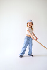 Young active girl in white crop top and wide jeans vigorously swings bat. She laughs and holds long stick in hands, stands in a dynamic pose on light background