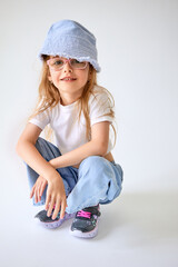 A seven-year-old girl in a white crop top,  jeans and a blue panama hat sits on her haunches. A child in glasses and sneakers with pink laces smiles on a cyclorama.