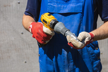A worker in a blue jumpsuit holds an electric screwdriver with a riveter attachment and an exhaust...